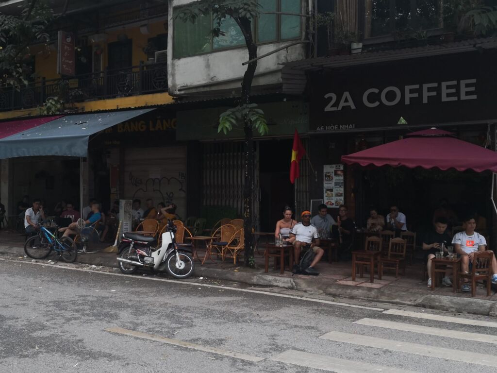 Strada con locali all'aperto biciclette e moto parcheggiati in strada, persone sedute ai tavoli bandiera piante sulle terrazze degli appartamenti soprastanti